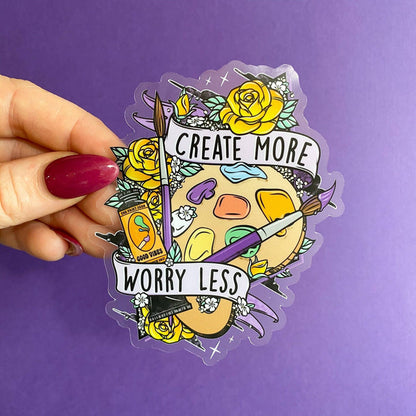 A transparent sticker with a colorful design, featuring text and imagery related to art creation, held against a purple background.