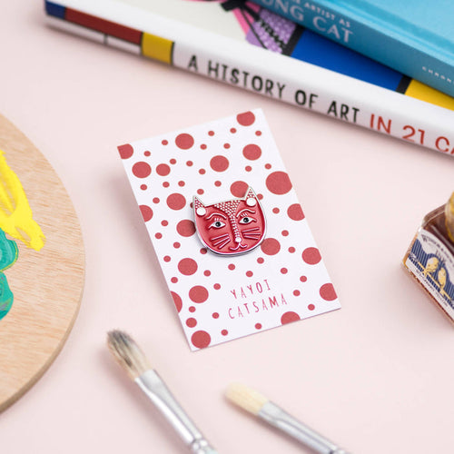 An enamel pin badge with a cat design, placed on a dotted card, with brushes and books in the background, suggesting an artistic theme.